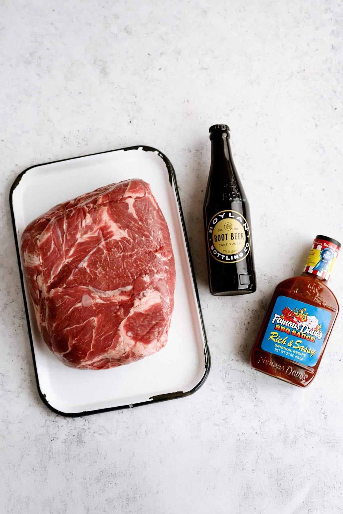 A raw beef roast on a white tray sits beside a bottle of root beer and a bottle of barbecue sauce on a light surface.