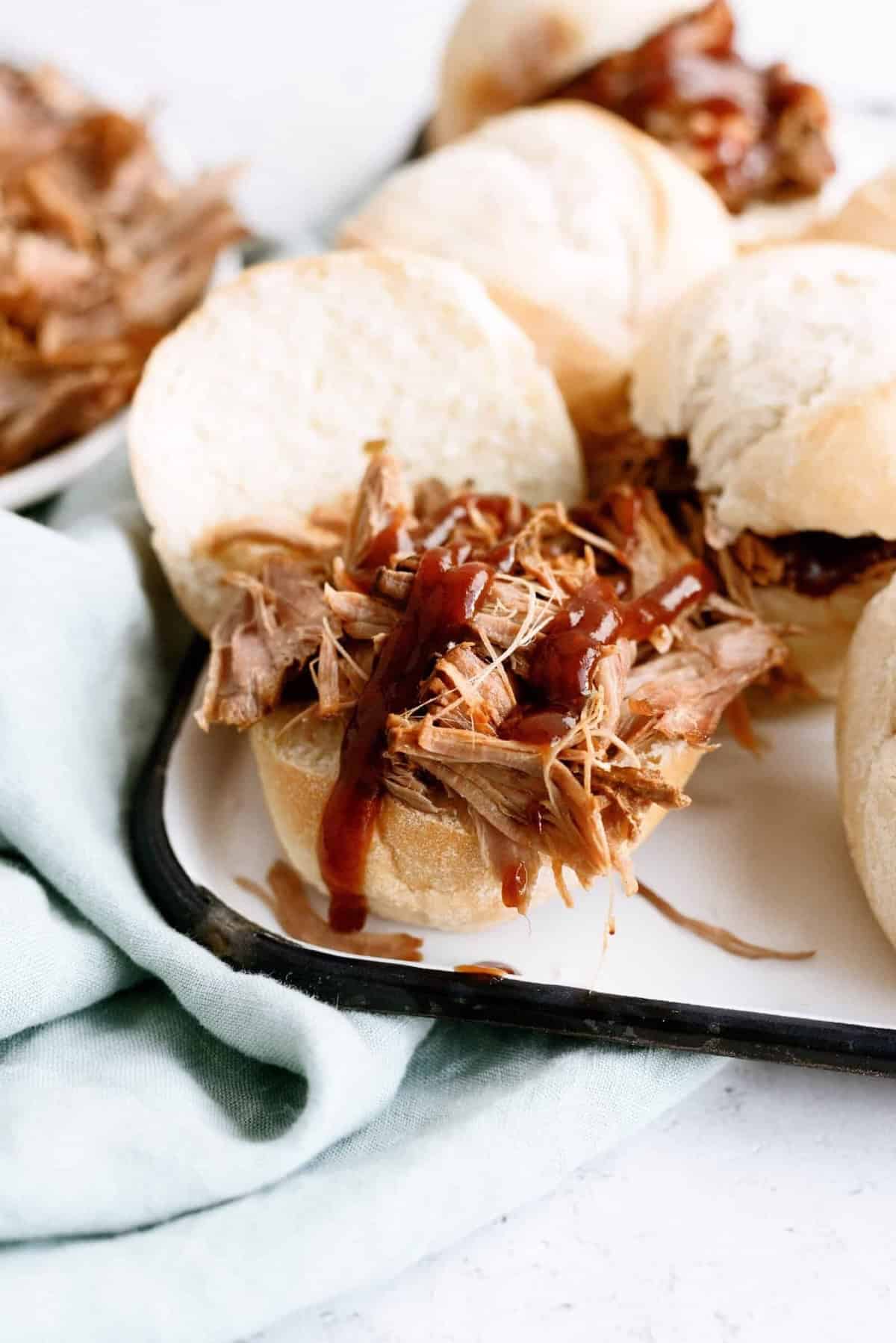 A shredded barbecue pork sandwich on a bun is served on a white plate with other sandwiches in the background.