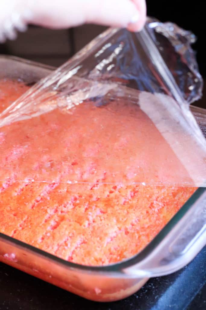 Strawberries and Cream Poke Cake in a glass pan with saran wrap being peeled off.