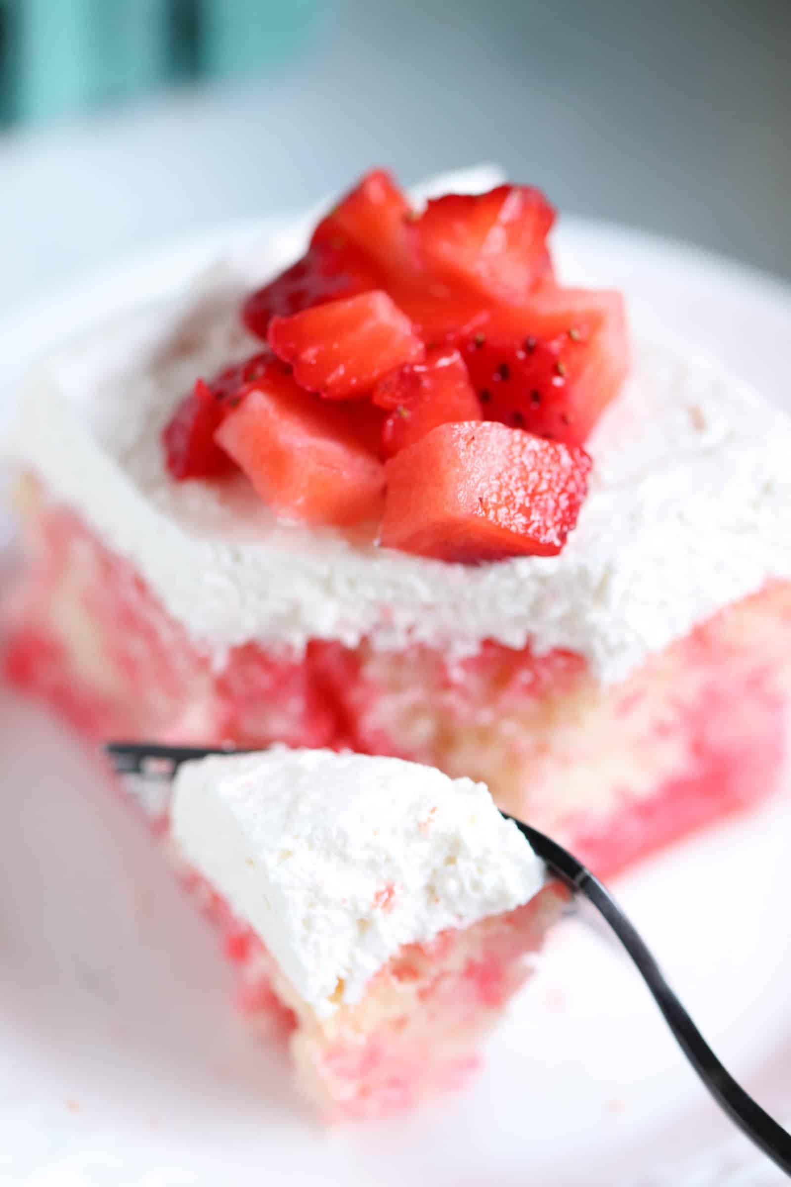 A slice of Strawberries and Cream Poke Cake on a plate with a fork taking a bite out of it.