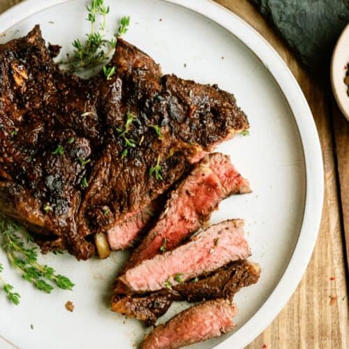 A cooked steak sliced to show a medium-rare center, garnished with fresh herbs, served on a white plate atop a wooden table.