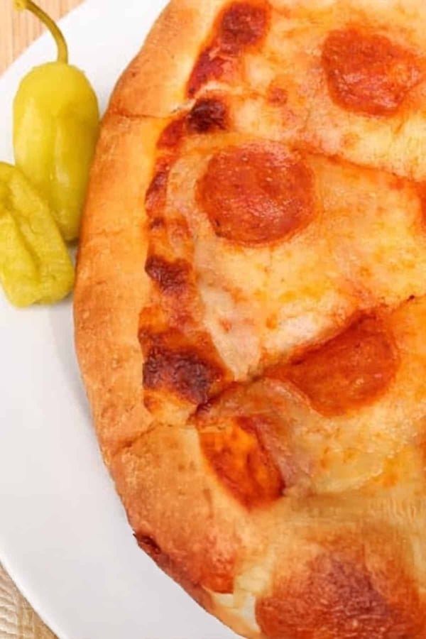 A close-up of a pepperoni pizza on a white plate with two yellow-green peppers on the side.