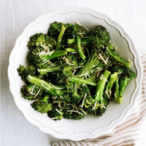 A white bowl filled with cooked broccoli florets topped with shredded cheese, placed on a white surface next to a striped cloth.