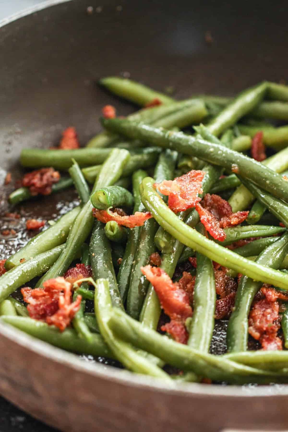 Green beans and crispy bacon pieces sautéing in a skillet.