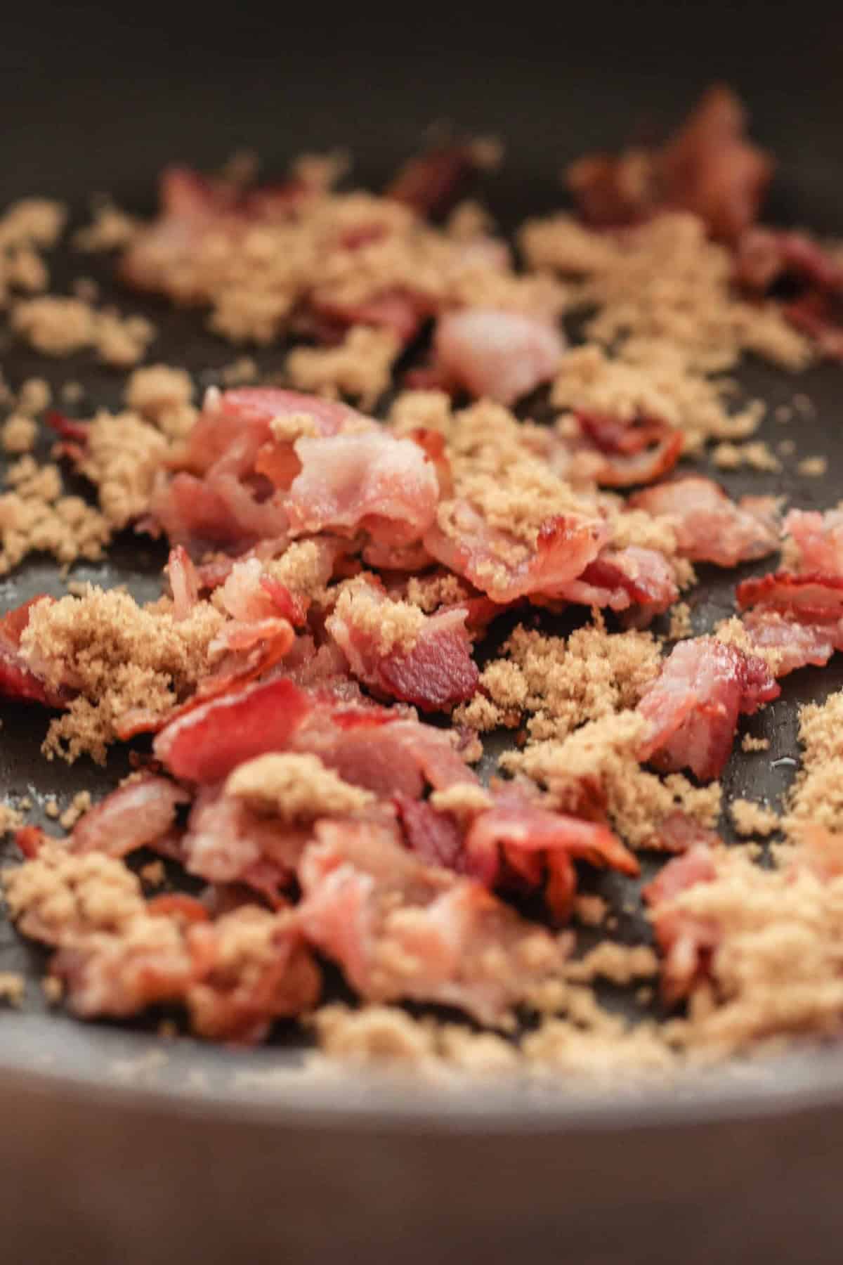 Crumbled cooked bacon pieces and brown sugar scattered in a frying pan.