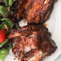 Two pieces of barbecued ribs served on a white plate with a side of mixed green salad and a cherry tomato.