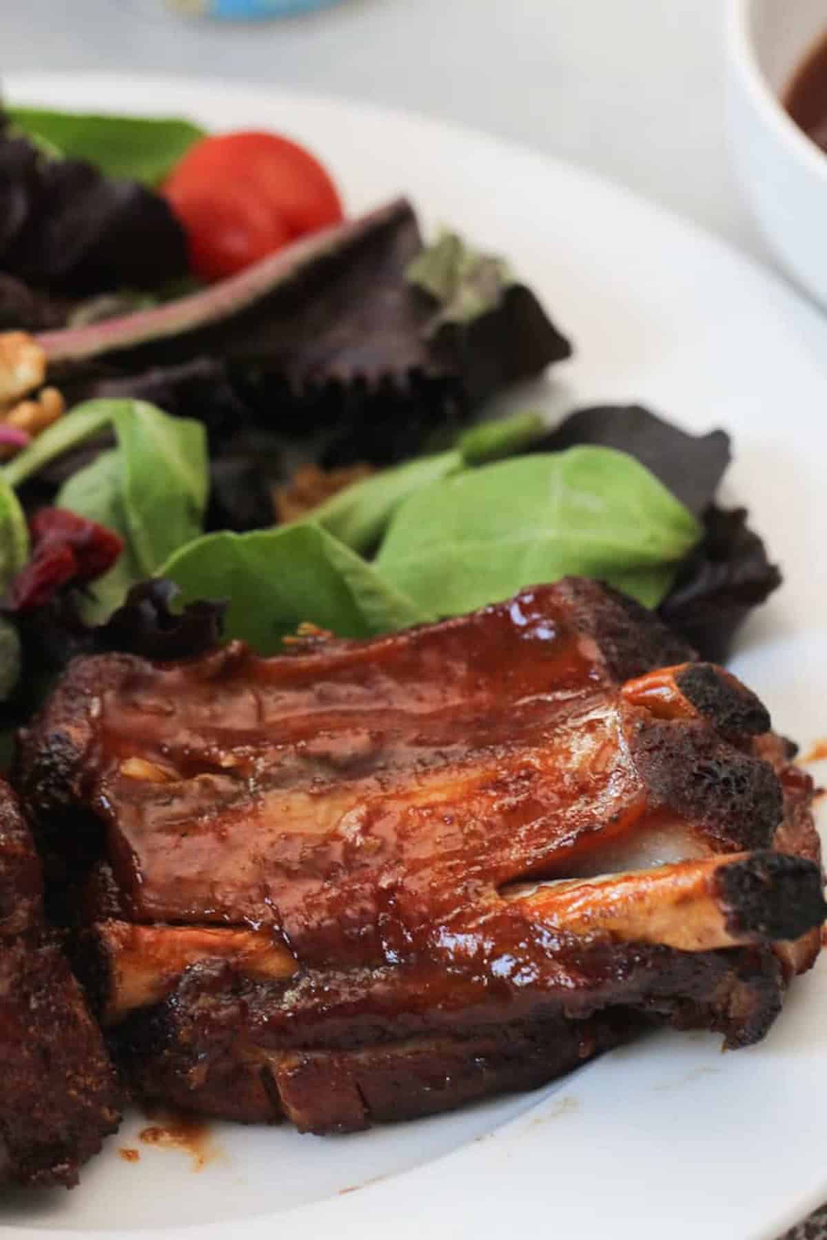 A plate with glazed barbecue ribs and a side of mixed green salad with cherry tomatoes.