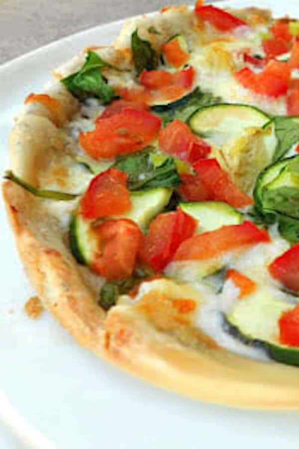 A close-up of a vegetable pizza topped with tomatoes, spinach, and zucchini on a white plate.