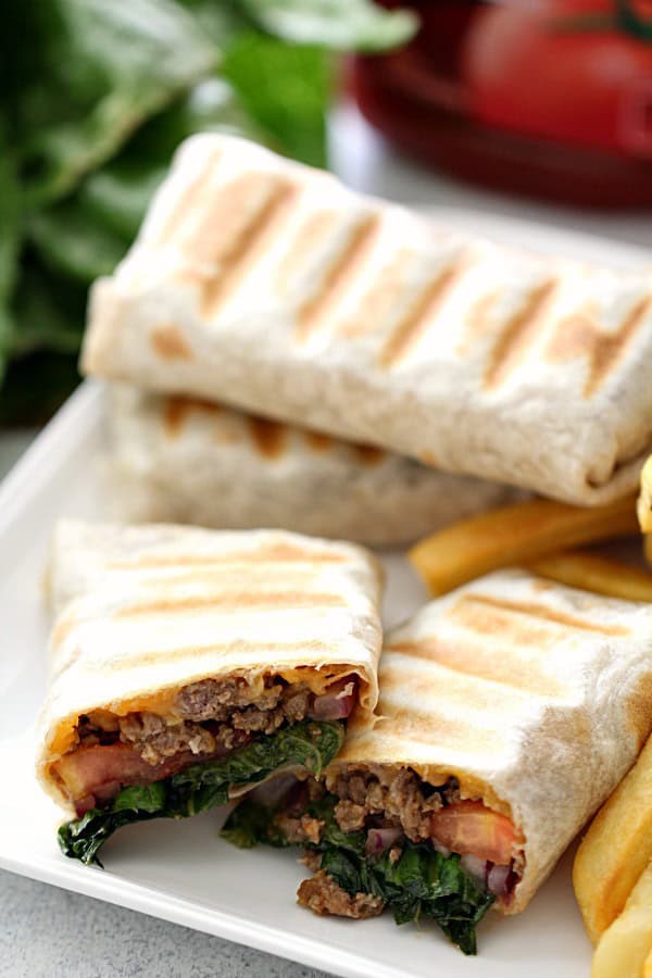 Close-up of two grilled wraps cut in half, revealing a filling of ground meat, lettuce, tomatoes, and cheese. A side of fries is visible on the plate. Background includes blurred leafy greens and tomatoes.