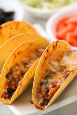 Three hard shell tacos filled with melted cheese and a mixture of beans and meat are arranged on a white plate, with bowls of diced tomatoes and other toppings in the background.