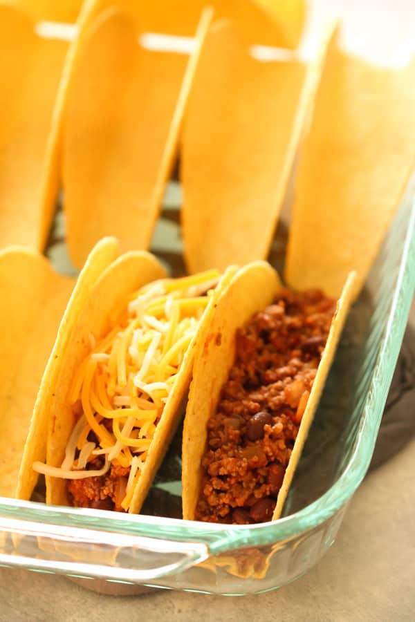 Oven Baked Beef Tacos in 9x13 dish with shredded cheese on top of one taco and just meat in the other shell