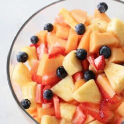 A glass bowl filled with a mixed fruit salad containing pineapple, cantaloupe, strawberries, and blueberries.
