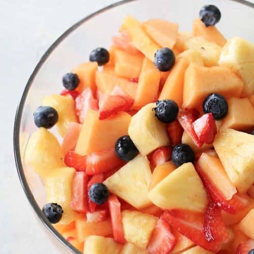 A glass bowl filled with a mixed fruit salad containing pineapple, cantaloupe, strawberries, and blueberries.