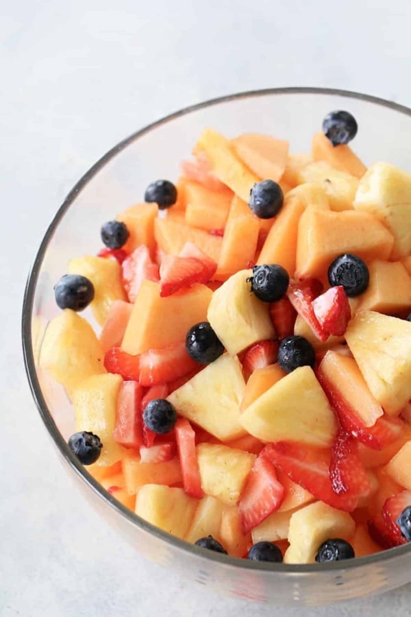 A glass bowl filled with a mixed fruit salad containing pineapple, cantaloupe, strawberries, and blueberries.