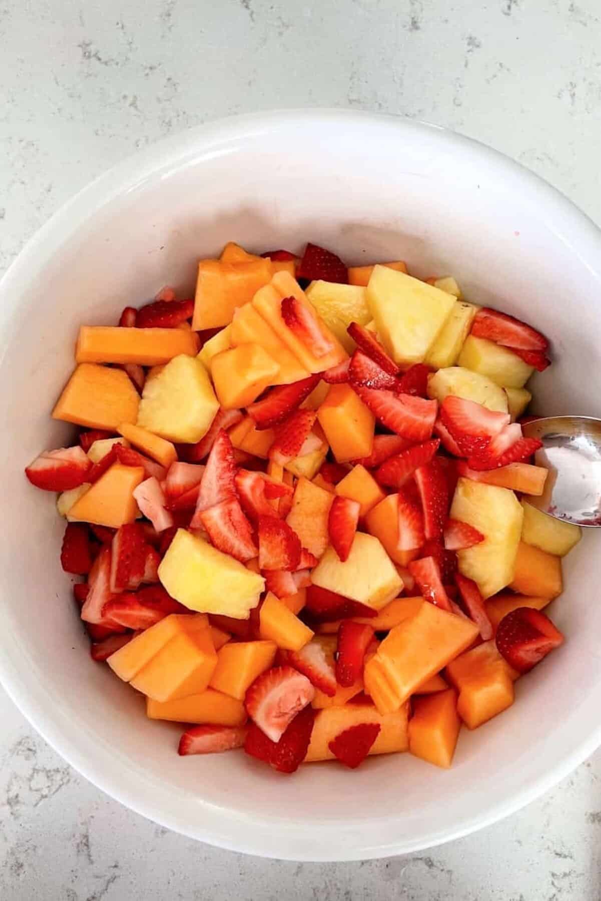 A white bowl filled with chopped strawberries, cantaloupe, and pineapple pieces, with a metal spoon on the side.