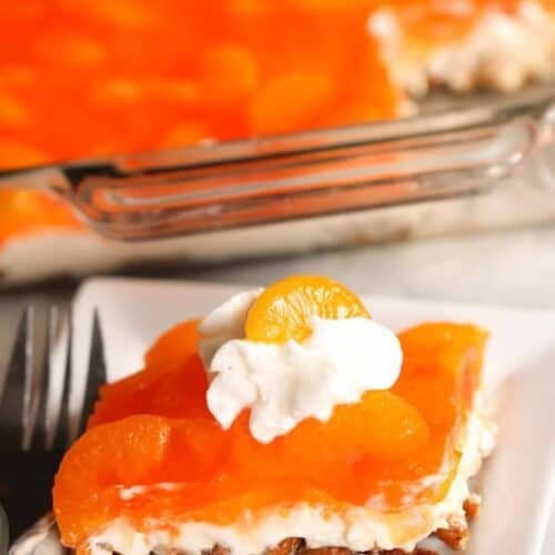 A slice of orange gelatin dessert with mandarin oranges, whipped cream, and a nutty crust on a white plate, with a fork beside it and a larger dish in the background.