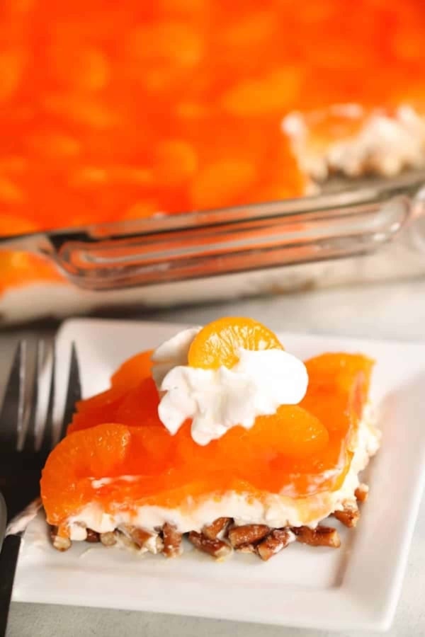 A slice of orange gelatin dessert with mandarin oranges, whipped cream, and a nutty crust on a white plate, with a fork beside it and a larger dish in the background.