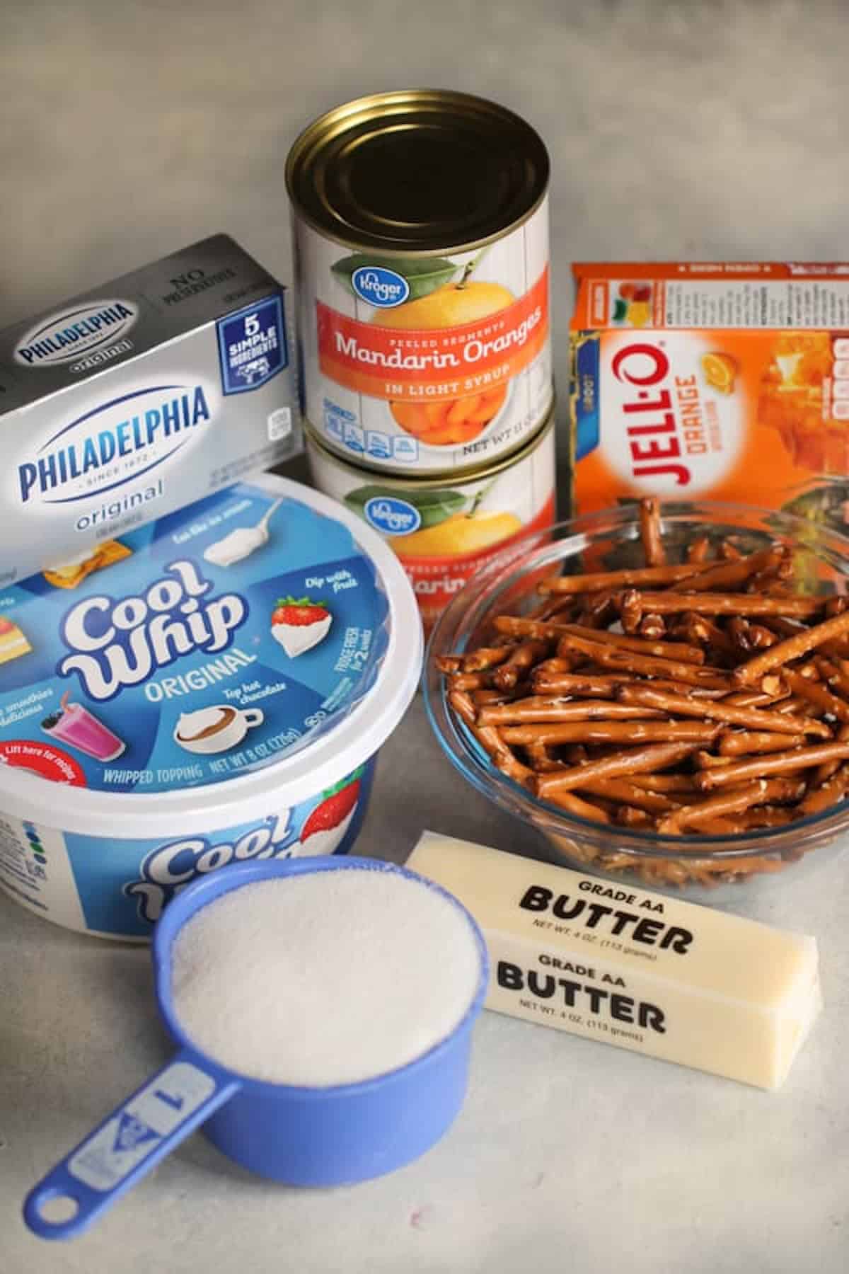 Ingredients for a dessert, including cream cheese, Cool Whip, mandarin oranges, orange Jell-O, pretzel sticks, a cup of sugar, and two sticks of butter, displayed on a counter.