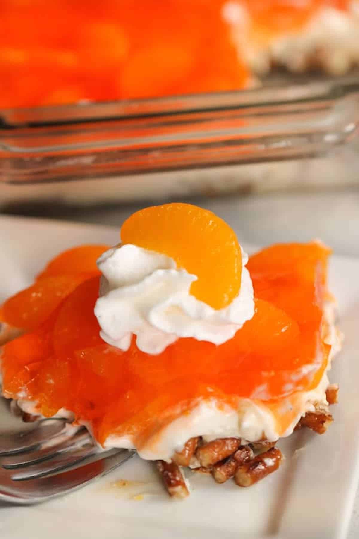A dessert bar with an orange gelatin topping, whipped cream, a mandarin orange slice, creamy layer, and a pecan crust on a white plate with a fork.