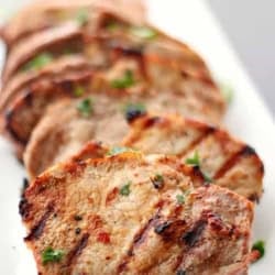Slices of grilled pork tenderloin garnished with chopped herbs are arranged on a rectangular white plate.
