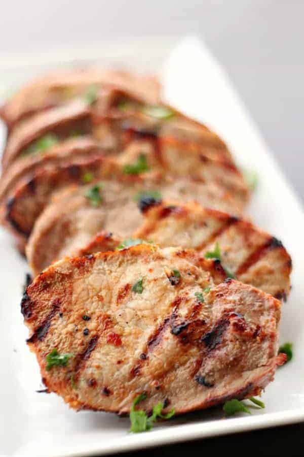 Slices of grilled pork tenderloin garnished with chopped herbs are arranged on a rectangular white plate.