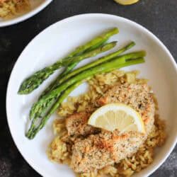 A white bowl with tilapia topped with a lemon slice, served over rice with three asparagus spears. A halved lemon is on the table beside the bowl.