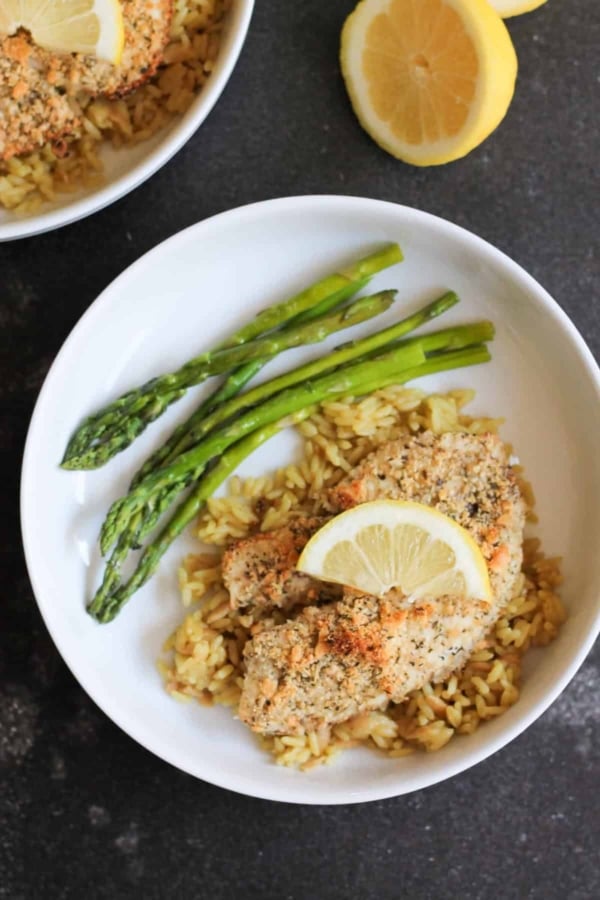 A white bowl with tilapia topped with a lemon slice, served over rice with three asparagus spears. A halved lemon is on the table beside the bowl.