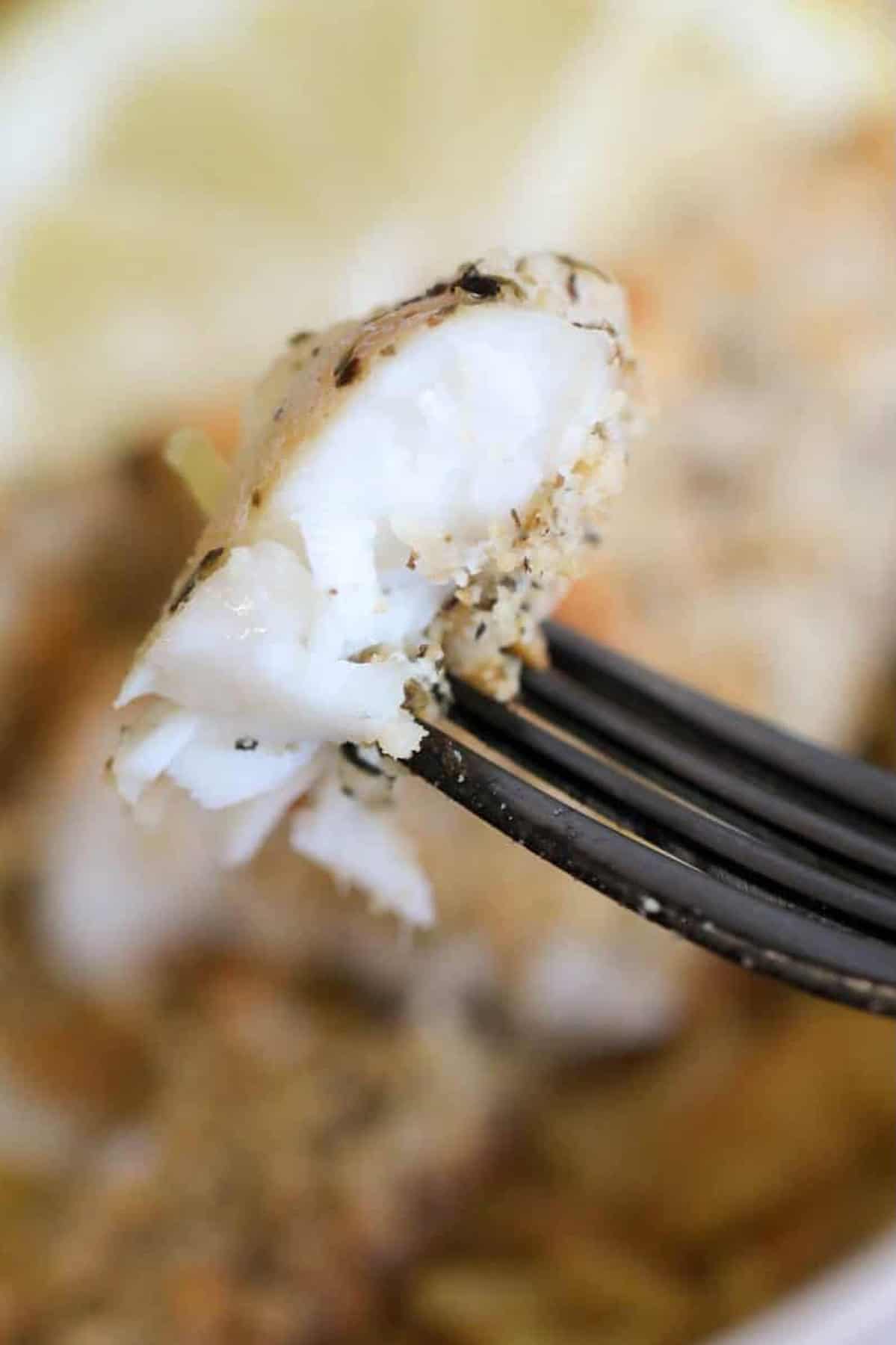 A close-up of a fork holding a piece of cooked white fish with herbs, with lemon slices blurred in the background.