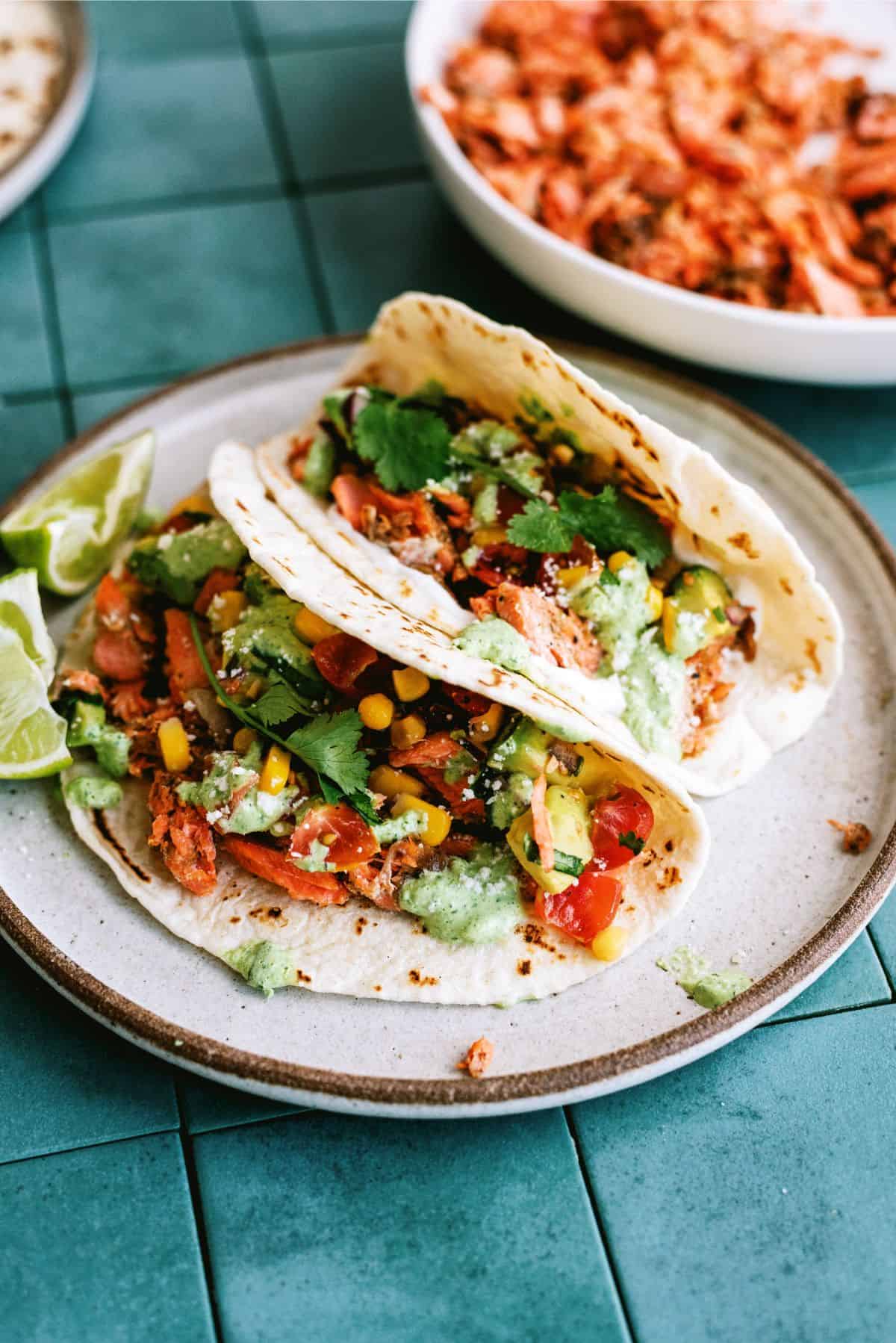 Two tacos filled with shredded salmon, corn, tomatoes, cilantro, and green sauce are served on a plate with lime wedges on a tiled surface.