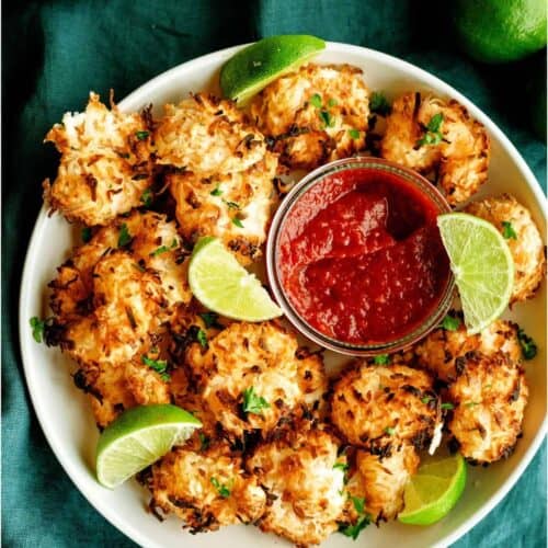 A plate of baked coconut shrimp garnished with lime wedges and parsley, served with a bowl of red dipping sauce on a green cloth.