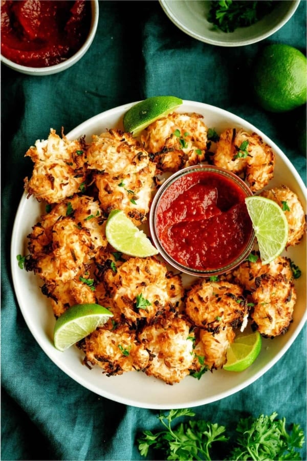 A plate of baked coconut shrimp garnished with lime wedges and parsley, served with a bowl of red dipping sauce on a green cloth.