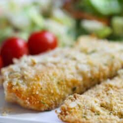 Two breaded fish fillets on a white plate with cherry tomatoes and a green salad in the background.