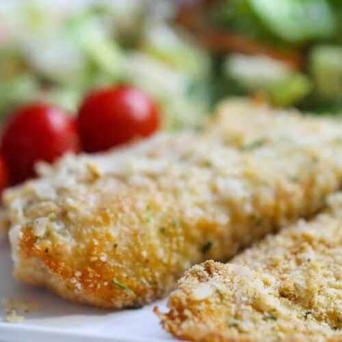 Two breaded fish fillets on a white plate with cherry tomatoes and a green salad in the background.