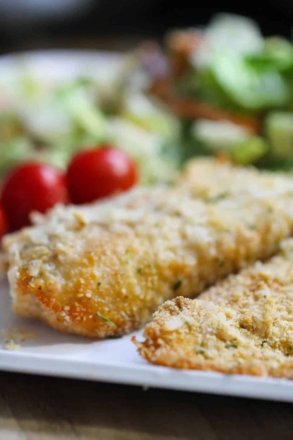 Two breaded fish fillets on a white plate with cherry tomatoes and a green salad in the background.