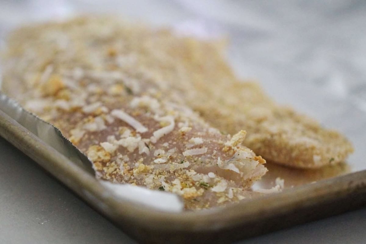 A close-up of a breaded fish fillet on a metal baking tray, ready for cooking.