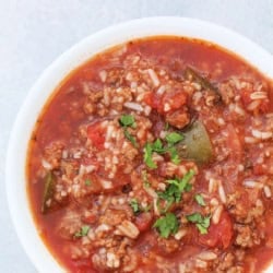 A white bowl filled with tomato-based soup containing ground meat, rice, chopped green peppers, and garnished with fresh herbs.