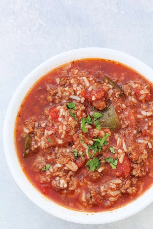A white bowl filled with tomato-based soup containing ground meat, rice, chopped green peppers, and garnished with fresh herbs.