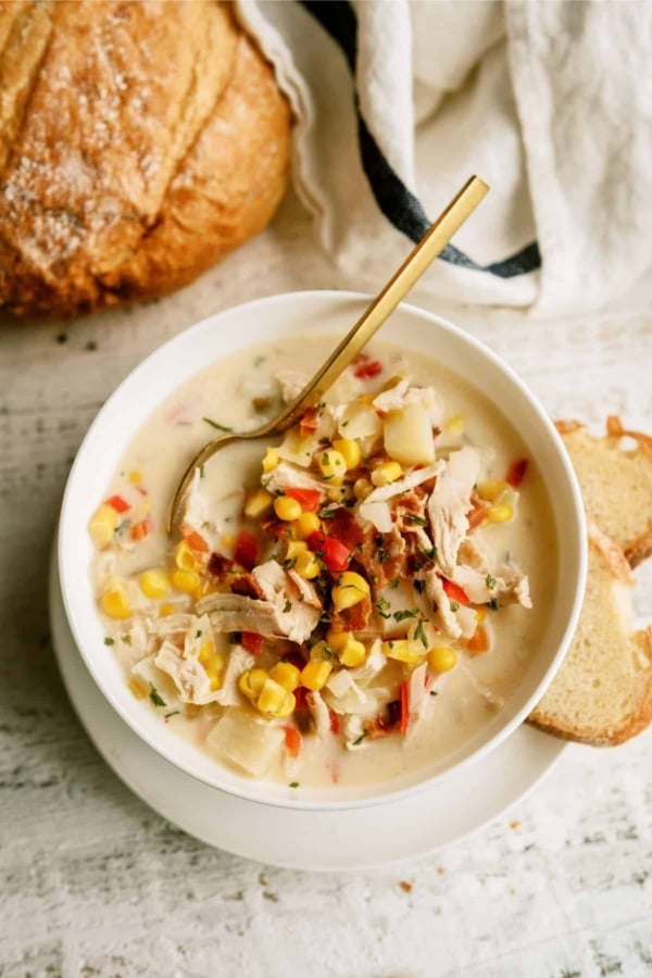 A bowl of creamy chicken and corn chowder garnished with herbs and bacon bits, accompanied by slices of bread on the side.