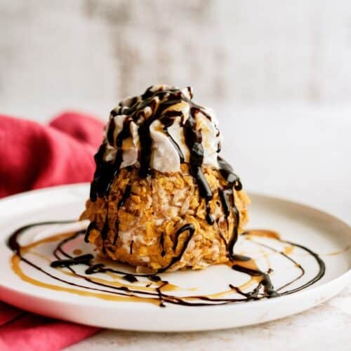 A scoop of fried ice cream topped with whipped cream and drizzled with chocolate and caramel sauce sits on a white plate.