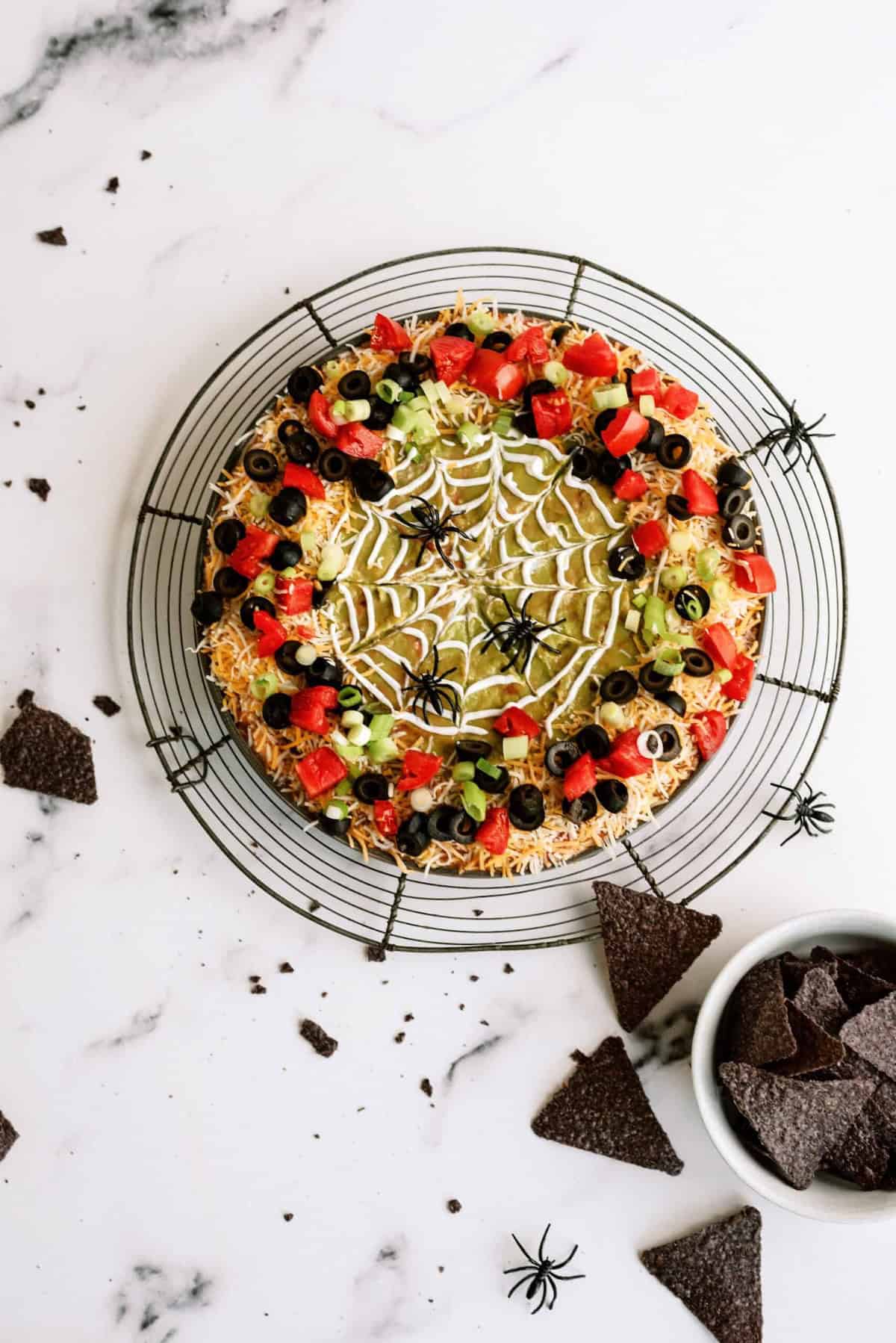 A round platter of layered dip decorated with a spider web-like topping and plastic spiders. Surrounding the platter are scattered chips and a bowl of tortilla chips.