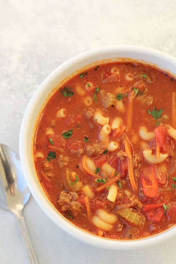 A close-up of a bowl of minestrone soup with visible macaroni, ground meat, chopped vegetables, and a tomato-based broth next to a spoon on a white surface.