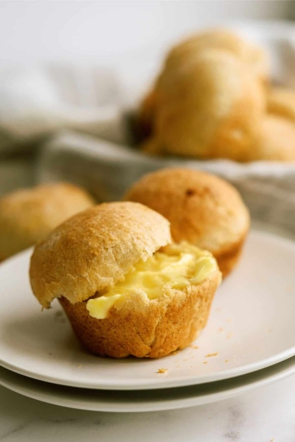 Two buttery rolls are placed on a white plate, one of which is split open, showing a generous amount of butter inside. More rolls are blurred in the background.