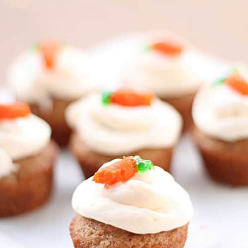 A close-up of mini cupcakes with white frosting and small orange and green decorations on top, arranged on a white plate.