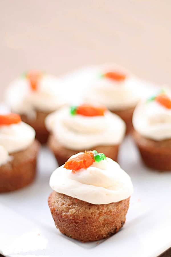 A close-up of mini cupcakes with white frosting and small orange and green decorations on top, arranged on a white plate.