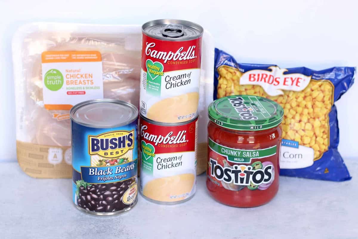 Assorted ingredients including raw chicken breasts, canned black beans, cream of chicken soup, chunky salsa, and a bag of frozen corn arranged on a white surface.