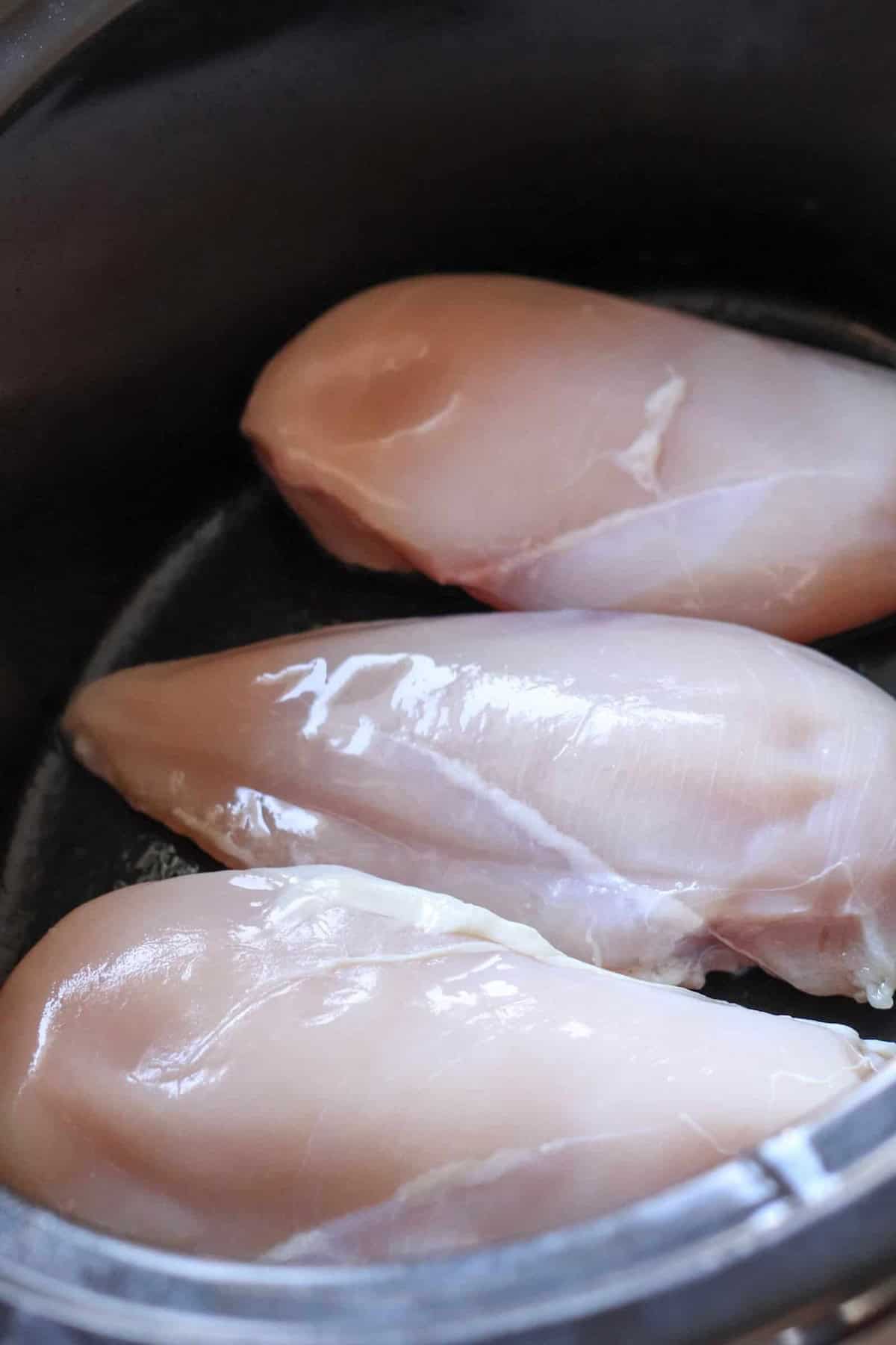 Three raw boneless, skinless chicken breasts placed in a slow cooker.