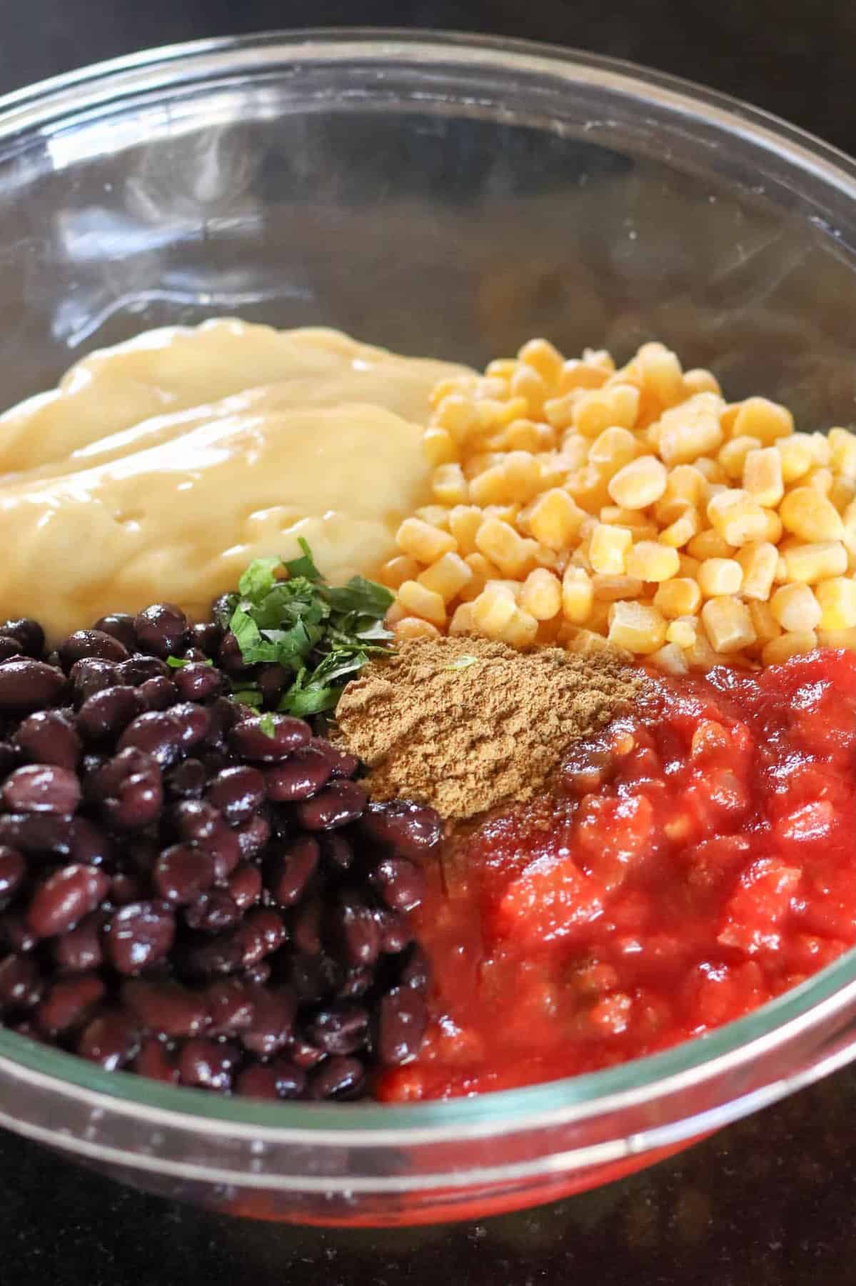 A glass bowl containing black beans, corn, salsa, cream of chicken soup, chopped herbs, and ground cumin, all separated into sections.