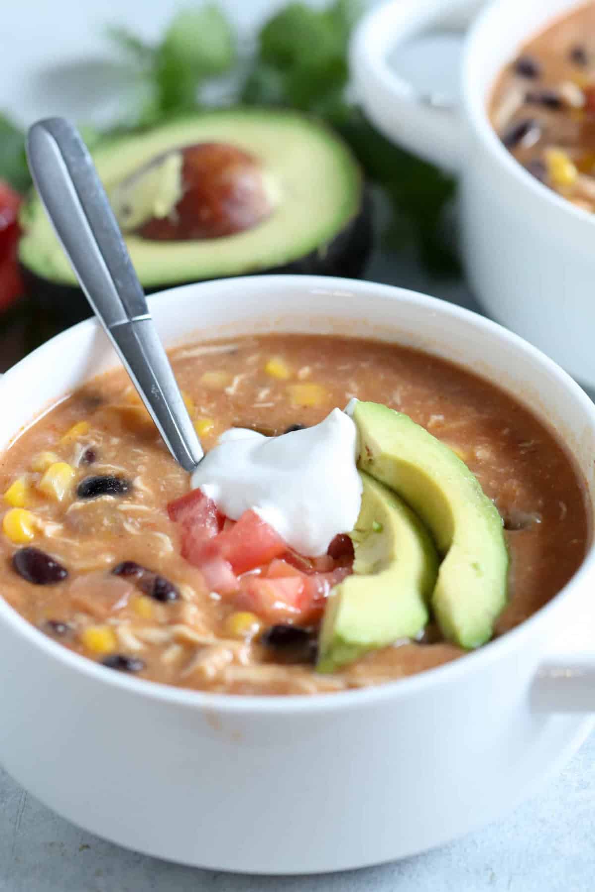 A bowl of creamy soup with black beans, corn, and chicken, topped with sliced avocado, diced tomatoes, and a dollop of sour cream, with a spoon in the bowl.
