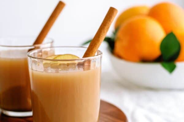 Two glasses of brown liquid with cinnamon sticks and a slice of lemon on a wooden tray; a bowl of oranges in the background.