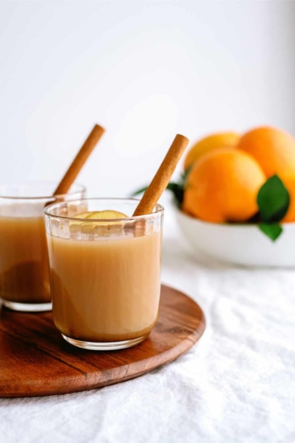 Two glasses of brown liquid with cinnamon sticks and a slice of lemon on a wooden tray; a bowl of oranges in the background.